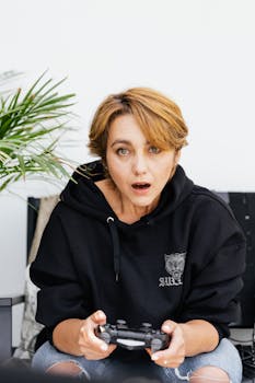 Woman in black hoodie playing video games, looking surprised indoors.
